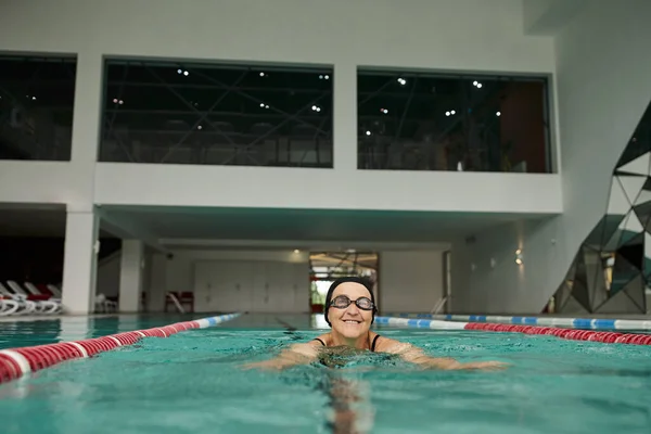 Mulher de meia-idade alegre em touca de natação e óculos nadando na piscina, centro de recreação, estilo de vida — Fotografia de Stock