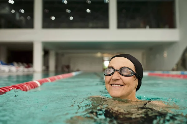 Mulher de meia-idade alegre em touca de natação e óculos nadando na piscina, água, centro de recreação, spa — Fotografia de Stock