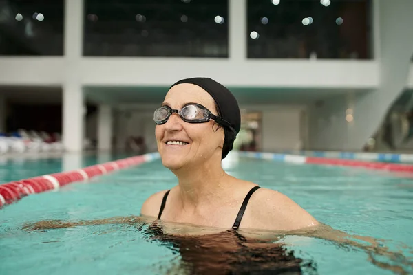 Позитивна жінка середнього віку в шапці для плавання та окулярах, що плавають у басейні, воді, центрі відпочинку, спа-центрі — стокове фото