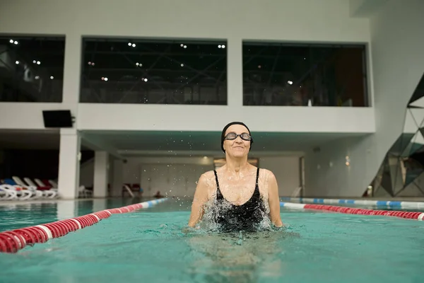 Salpicos de água, mulher de meia idade em touca de natação e óculos, sorrindo, exercitando-se dentro da piscina — Fotografia de Stock