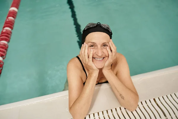 Top view of happy middle aged woman in swim cap and goggles, water in pool, recreation center — Stock Photo