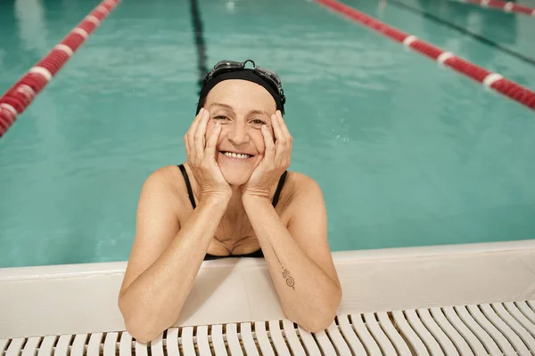 Joyous middle aged woman in swim cap and goggles looking at camera, water in pool, recreation center — Stock Photo