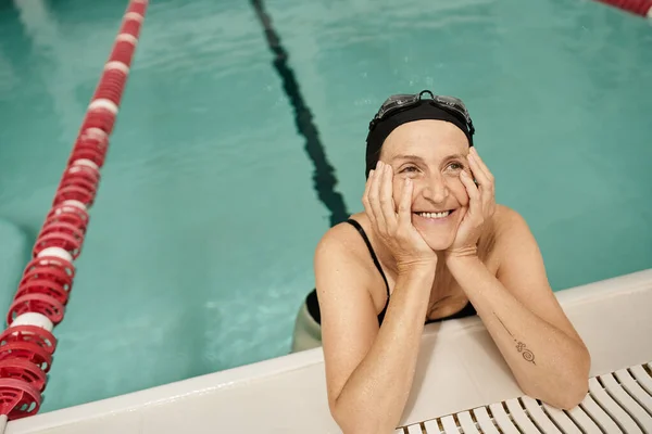 Middle aged woman in swim cap and goggles looking away, smiling, water in pool, recreation center — Stock Photo