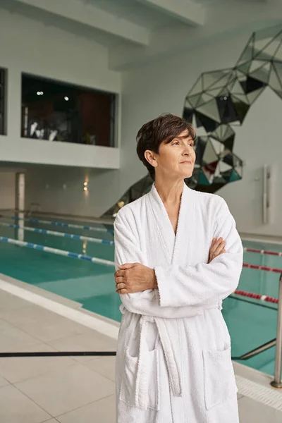 Mulher de meia idade com cabelo curto em pé com braços dobrados, vestindo roupão branco perto da piscina, dia spa — Fotografia de Stock