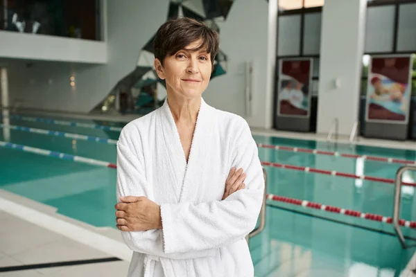 Donna di mezza età con i capelli corti in piedi con le braccia piegate vicino alla piscina, accappatoio bianco, centro benessere — Foto stock