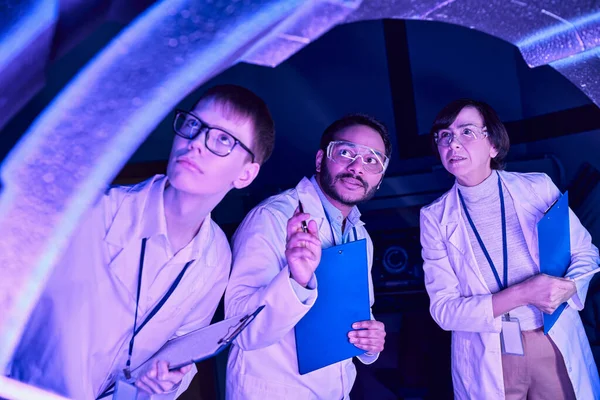 Futuristische Exploration: Wissenschaftler verschiedener Zeitalter untersuchen Gerät im Neon-Lit Science Center — Stockfoto