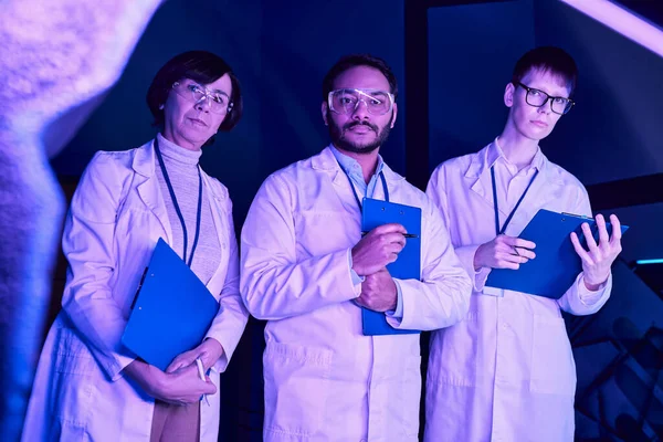 Observación futurista: Tres científicos examinan un dispositivo recién creado en el Neon-Lit Science Center - foto de stock