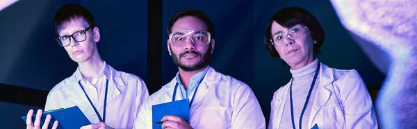 Banner, Tres científicos examinan dispositivo recién creado en Neon-Lit Science Center - foto de stock