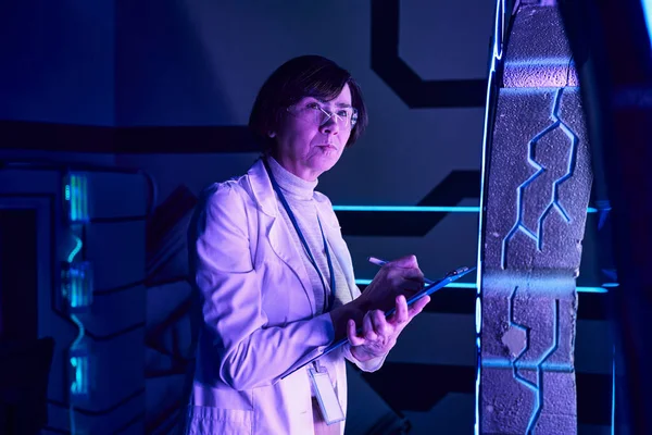 Futuristic concept, thoughtful woman scientist writing on clipboard in neon-lit discovery center — Stock Photo