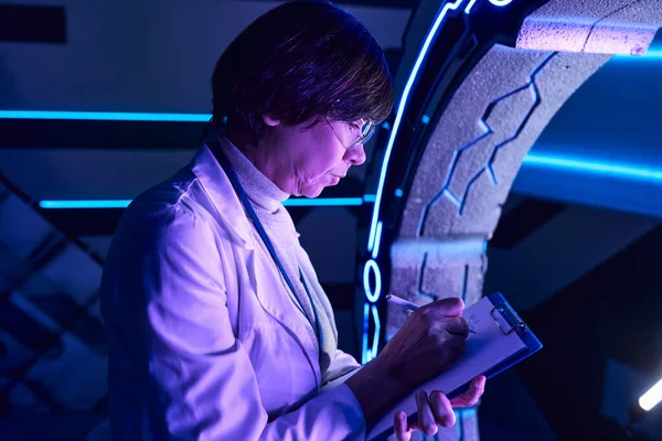 Pesquisa futurista, experiente mulher cientista escrevendo em prancheta perto de novos equipamentos inovadores — Fotografia de Stock