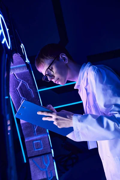 Young intern writing on clipboard near new experimental equipment in science center of future — Stock Photo