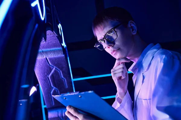 Young thoughtful scientist with clipboard in neon-lit innovation hub, futuristic concept — Stock Photo