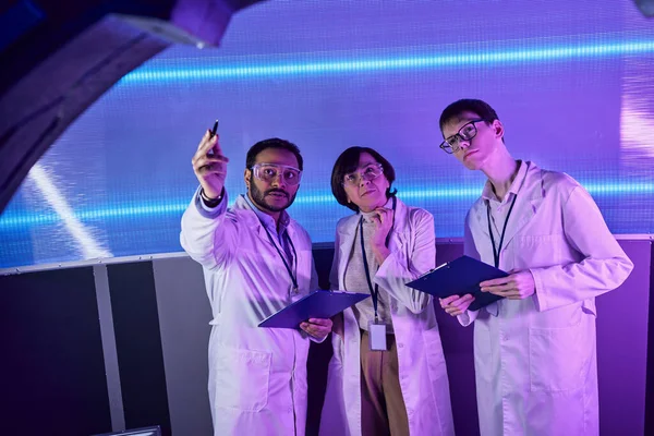 Scientifique indien pointant avec stylo vers le nouvel appareil près de collègues dans le centre de découverte futuriste — Photo de stock
