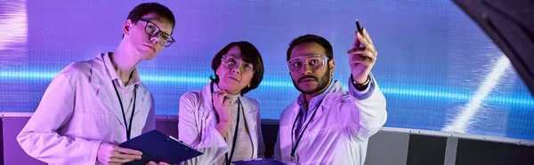 Uomo indiano che punta con penna vicino agli scienziati nel centro scientifico futuristico illuminato al neon, striscione — Foto stock