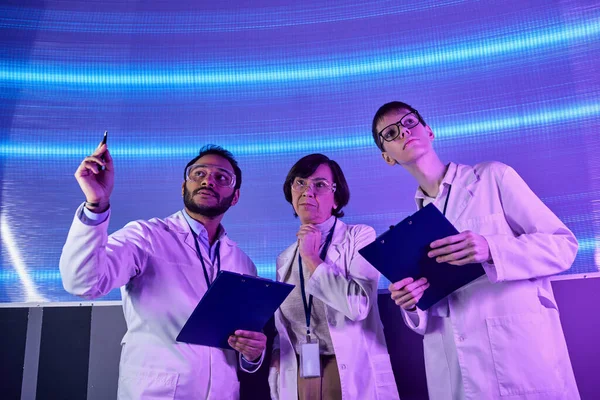 Future-oriented, indian scientist pointing with pen near team in innovative science center — Stock Photo