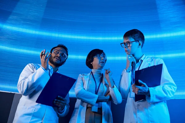 Soluciones futuristas, científicos multiétnicos con portapapeles hablando en el centro de descubrimiento con luz de neón - foto de stock