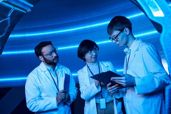 Cientistas multiétnicos olhando para o jovem estagiário com área de transferência no centro de ciência futurista iluminado por néon — Fotografia de Stock