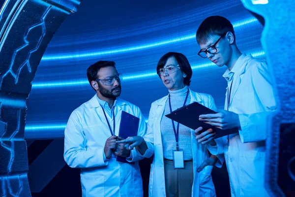 Ученый средних лет указывает на экспериментальное устройство рядом с многонациональными коллегами в научном центре — стоковое фото