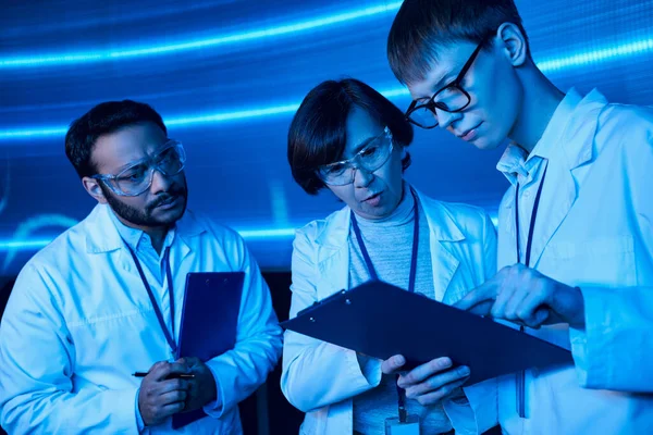 Junge Praktikantin zeigt auf Klemmbrett in der Nähe multiethnischer Kollegen im Science Center, zukunftsorientiert — Stockfoto