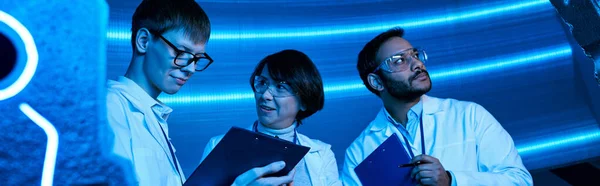 Young intern looking at clipboard near multiethnic colleagues in neon-lit science center, banner — Stock Photo