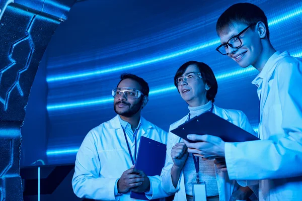 Young intern with clipboard smiling near multiethnic colleagues in futuristic science center — Stock Photo