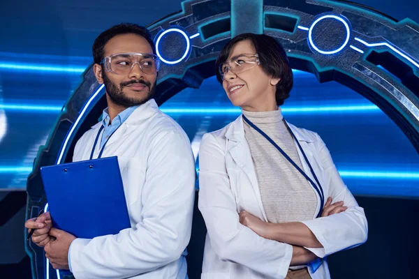Laboratório inovador, cientistas multiétnicos sorrindo uns para os outros perto de dispositivo experimental iluminado por néon — Fotografia de Stock