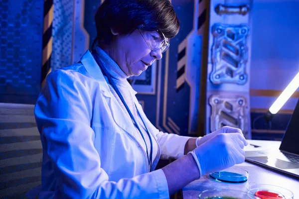 Женщина среднего возраста в очках работает с образцами инопланетной жизни в чашках Петри в лаборатории — стоковое фото