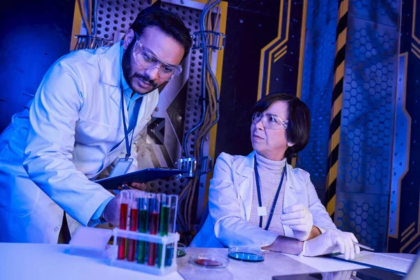 Científico indio mirando tubos de ensayo con muestras de vida extraterrestre cerca de colega en el laboratorio iluminado por neón - foto de stock