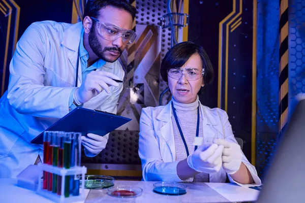 Indian scientist pointing with pan at samples near colleague in futuristic scientific laboratory — Stock Photo