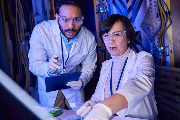 Indian scientist with clipboard pointing at laboratory glass near colleague in innovative laboratory — Stock Photo