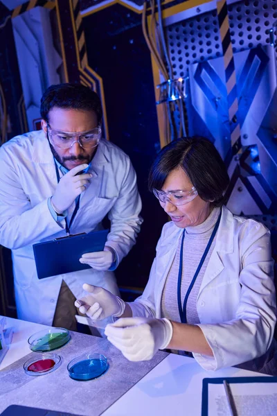 Científico indio con portapapeles mirando vidrio de laboratorio cerca de colegas y placas de Petri - foto de stock
