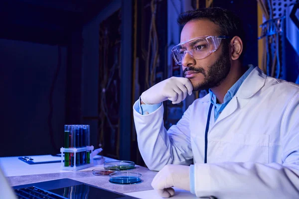 Indian scientist in goggles working on laptop near test tubes and petri dishes in futuristic lab — Stock Photo
