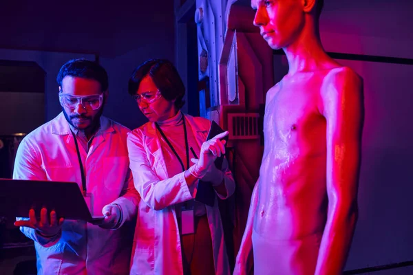 Wissenschaftlerin zeigt mit Laptop in Innovationszentrum auf humanoiden Außerirdischen in der Nähe indischer Kollegen — Stockfoto