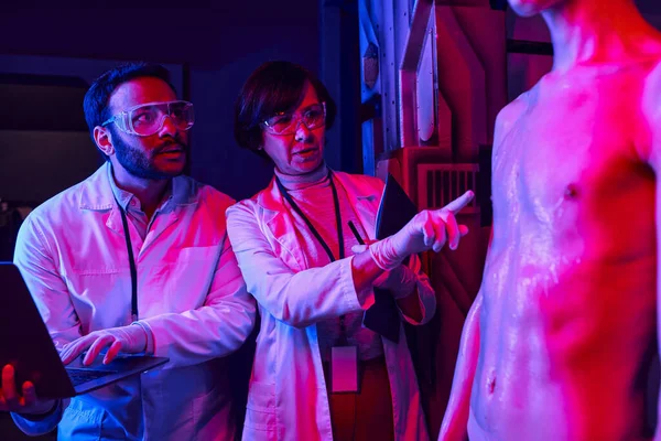 Científico indio con portátil cerca de colega señalando desconocido humanoide extranjero en el centro de la ciencia - foto de stock