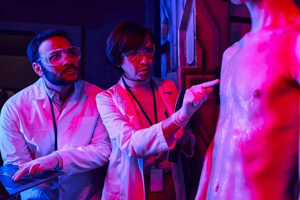Wissenschaftlerin berührt außerirdisches Wesen nahe indischem Kollegen mit Laptop im Labor — Stockfoto