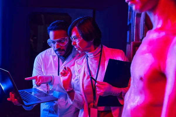 Indian scientist pointing at laptop near colleague and humanoid alien in science center — Stock Photo