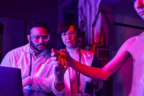 Científico indio con portátil cerca colega cogido de la mano de extraterrestre cósmico en laboratorio innovador - foto de stock