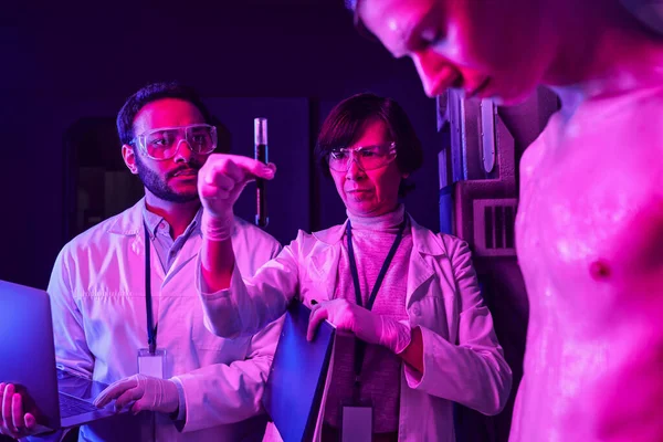Científico sosteniendo tubo de ensayo con muestra de sangre cerca de colega indio con portátil y extranjero en el laboratorio - foto de stock