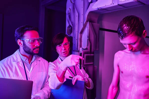 Científicos multiétnicos mirando el tubo de ensayo con muestra de sangre cerca de humanoides alienígenas en el centro de la ciencia - foto de stock