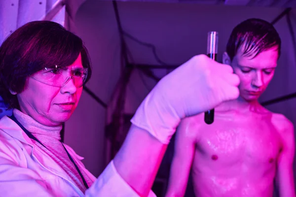 Ученый средних лет в очках смотрит на пробирку с образцом крови рядом с космическим существом в лаборатории — стоковое фото