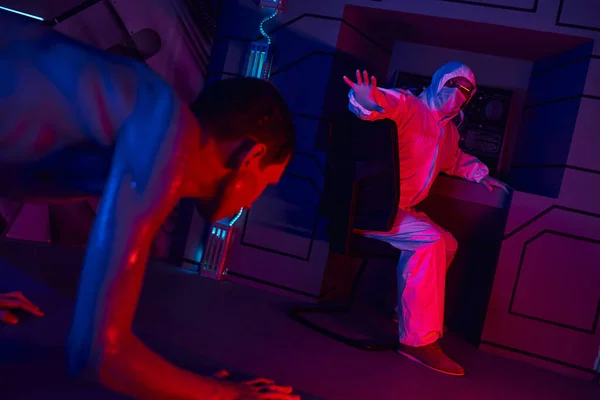 Humanoide extraterrestre arrastrándose cerca de un científico en traje de materiales peligrosos mostrando un gesto de parada en el laboratorio - foto de stock