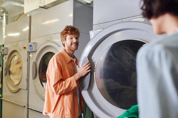 Homem sorridente em pé perto da máquina de lavar roupa e namorada borrada com bacia na lavanderia auto-serviço — Fotografia de Stock