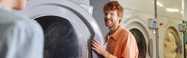 Giovane uomo sorridente in piedi vicino a lavatrici e fidanzata in lavanderia self service, banner — Foto stock
