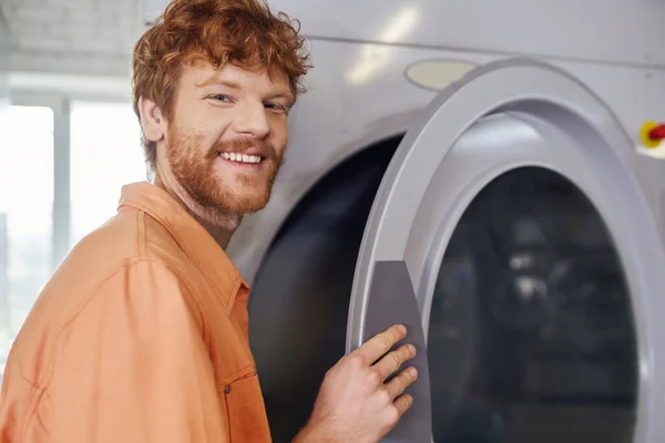 Усміхнений молодий рудий чоловік дивиться на камеру біля пральної машини в пральні самообслуговування — стокове фото