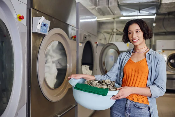 Усміхнена молода азіатка тримає кошик з одягом біля пральної машини в монетному пральні — стокове фото
