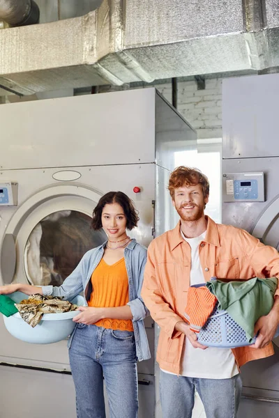 Sonriente pareja joven multiétnica sosteniendo lavabos con ropa cerca de la lavadora en la lavandería de monedas - foto de stock
