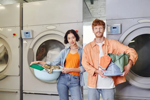 Sorrindo casal interracial segurando bacias perto de máquinas de lavar roupa na lavanderia moeda — Fotografia de Stock