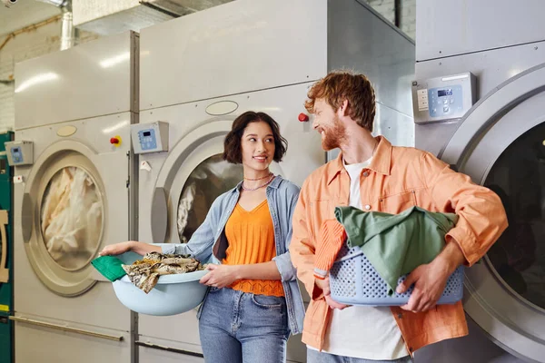 Casal interracial positivo segurando roupas perto de máquinas de lavar roupa na lavanderia moeda — Fotografia de Stock