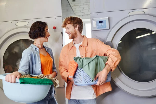 Sorrindo jovem casal multiétnico segurando bacias com roupas em lavanderia self-service — Fotografia de Stock