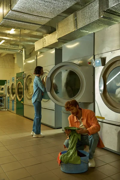 Young redhead man holding clothes near basin and asian girlfriend in coin laundry — Stock Photo